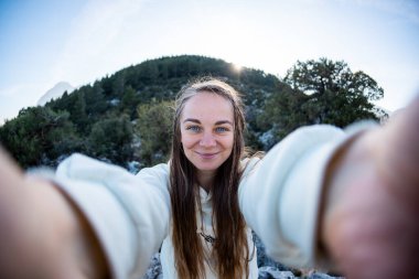 Young woman with blond hair taking a selfie portrait while hiking in the mountains - Happy hiker on top of a cliff smiling at camera - Travel and hobby concept