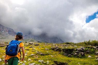 Sırt çantalı bir çocuk dağların arka planında yürüyüş yapıyor. Sırt çantalı çocuk gezgini, yürüyüş, seyahat, yaşam tarzı konsepti, arka planda dağlar, yaz tatili çocukları..