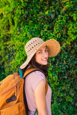 Yeşil bir duvarın arkasında sırt çantalı ve şapkalı genç bir kızın portresi. Yaz tatili. tatil ve macera konsepti.