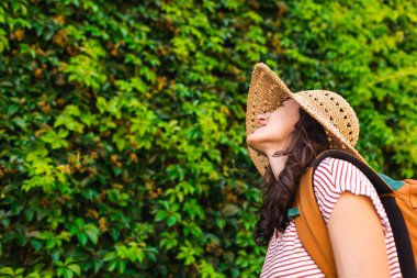 Yeşil bir duvarın arkasında sırt çantalı ve şapkalı genç bir kızın portresi. Yaz tatili. tatil ve macera konsepti.