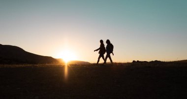 silhouette of two girls with backpacks while traveling in the mountains at sunset. two people walk along the mountain range at sunset. travel with freedom.