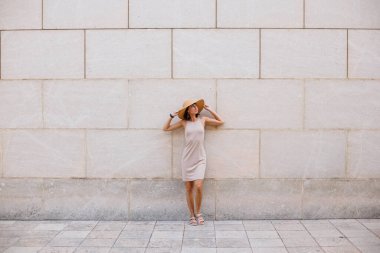 Moda bir yaz elbisesi içinde gülümseyen güzel bir kız. Sokakta şapkalı, duvara karşı poz veren seksi kaygısız kadın. Eğlence ve kahkaha.