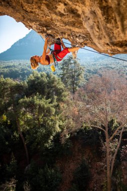 Kız kaya tırmanışçısı bir ormanın arka planına ve mavi dağlara tırmanır. Kaya tırmanışçısı zor bir yolda dinleniyor. Türkiye 'de kaya tırmanışı.