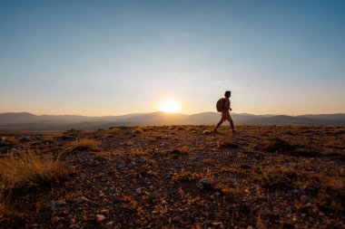 Sırt çantalı bir adam dağların arka planında yol boyunca yürür. Sırt çantasıyla dağlarda yürüyüş. Yürüyüş ve seyahat kavramı.