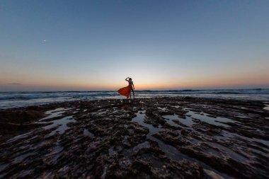Gün batımında deniz kenarında poz veren kırmızı uzun elbiseli güzel, şehvetli bir kadın. Güzel gün batımı. Havada uçan bir kumaş şeridi..