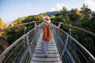 Güneşli bir sonbahar gününde, nehri geçen yaya asma köprüsü boyunca şapkalı mutlu bir kadın yürüyor. arka plan.