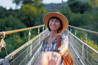 Bir nehrin üzerindeki asma köprüde yürüyen bir kadının sonbahar dönemi açık hava portresi. Kız döndü ve sanki beni takip ediyormuş gibi kameraya elini uzattı. Seyahat ve macera.