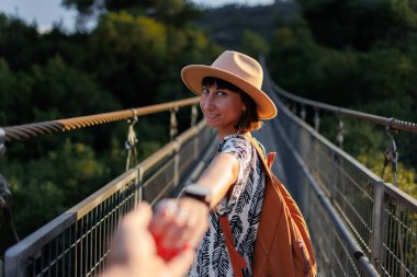 Bir nehrin üzerindeki asma köprüde yürüyen bir kadının sonbahar dönemi açık hava portresi. Kız döndü ve sanki beni takip ediyormuş gibi kameraya elini uzattı. Seyahat ve macera.