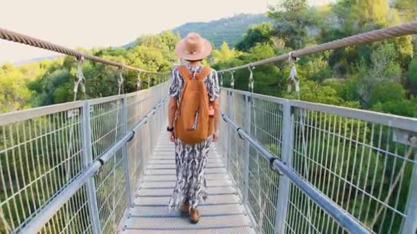 一位女游客走过河上的一座桥 一位年轻女子在秋天的森林前 沿着一条横跨河流的悬索桥行走 戴帽子的女孩 — 图库视频影像