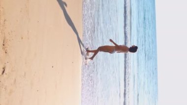 Dikey çekim. Güzel bir kız gün batımında bikinisiyle sahilde yürüyor, tatili boyunca deniz ve güneşin tadını çıkarıyor..