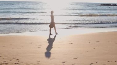 Elinde şapkası olan güzel bir kız gün batımında beyaz bir bikiniyle sahilde yürüyor. Tatili boyunca deniz ve güneşin tadını çıkarıyor..