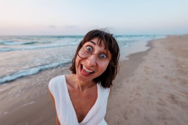 Beyaz elbiseli güzel, gülümseyen bir esmer, denizin arka planında kameraya mutlu bir şekilde bakar. Genç bir kızın portresi.