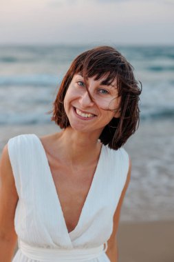 Beyaz elbiseli güzel, gülümseyen bir esmer, denizin arka planında kameraya mutlu bir şekilde bakar. Genç bir kızın portresi.