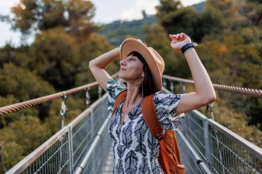 Sonbahar havası. Bir kadın turist nehir boyunca köprü boyunca yürür. Genç bir kadın, sonbahar ormanının önündeki bir nehrin karşısındaki asma köprü boyunca yürüyor. Şapkalı kız..