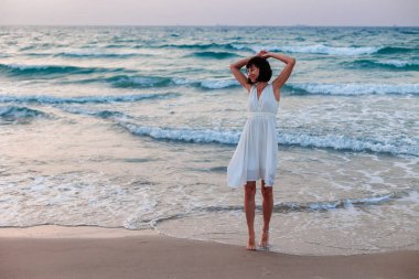 Beyaz elbiseli güzel, gülümseyen bir esmer, denizin arka planında kameraya mutlu bir şekilde bakar. Genç bir kızın portresi.