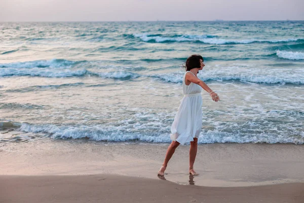 Happy traveler girl in a white summer dress enjoying a tropical paradise beach with turquoise sea. Happy young woman in a beautiful white dress walks along the beach during sunset.