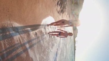 Dikey çekim. Mutlu genç anne ve oğlu deniz sahilinde yürüyor, gülüyor ve birlikte eğleniyorlar. Çocuğu olan genç bir anne. Aile deniz tatili.