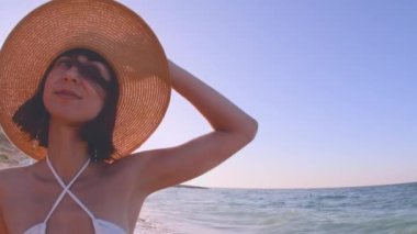 Sahildeki genç kız. Saman şapkalı ve deniz kıyısında beyaz bikinili güzel bir genç kadının portresi. Siyah saçlı mutlu kız güneşin tadını çıkarıyor..