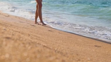 Kız sahilde yavaşça yürüyor ve dalgaların ve denizin tadını çıkarıyor. Bacaklar yakın. Mutlu genç kız gün batımında sahilde yürüyor..