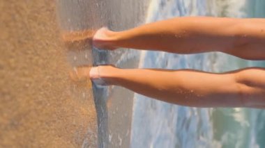 Dikey çekim. Bir kız sahilde duruyor ve dalgaların ve denizin tadını çıkarıyor. Bacaklar yakın. Kumsalda duran mutlu genç kız.