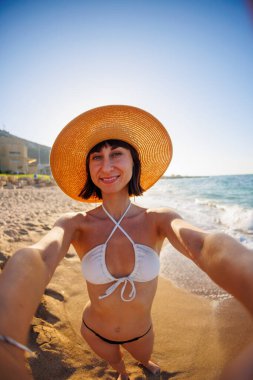 Sahilde selfie çeken, yaz şapkalı neşeli bir genç kızın yakın çekimi. Bir kız sahilde tatil yaparken selfie çekiyor..