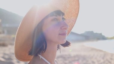 Deniz kıyısında hasır şapkalı güzel bir genç kadının portresi. Yaz tatili boyunca sahilde gülümseyen neşeli genç bir kadın. Siyah saçlı mutlu kız güneşin tadını çıkarıyor..