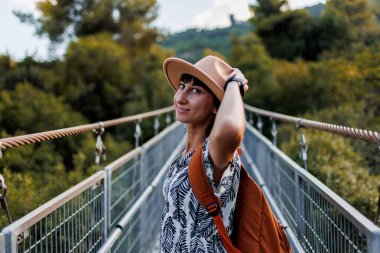 Sonbahar havası. Bir kadın turist nehir boyunca köprü boyunca yürür. Genç bir kadın, sonbahar ormanının önündeki bir nehrin karşısındaki asma köprü boyunca yürüyor. Şapkalı kız..