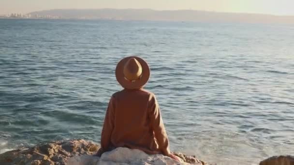 后视镜一个头戴帽子 身穿外套的轻松自在的女孩坐在海边的石头上 一个女孩坐在岩石上看着大海 — 图库视频影像