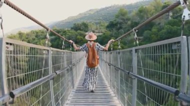 Sırt çantasıyla seyahat ediyor. Şapkalı ve sırt çantalı genç bir kız nehir boyunca bir köprü boyunca yürüyor. Şapkalı kız. seyahat ve macera.
