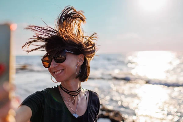 Close-up of a stunning woman with hair blowing in the wind taking a selfie by the sea. Beautiful girl smiling while looking at camera outside - fashionable lifestyle and beauty concept