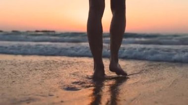 Gün batımında bir kız kumsalda yalınayak yürüyor, kumda ayak izleri ve suda yansımalar bırakıyor. Sahilde kadın bacaklarına yakın çekim. Sakinlik ve sükunet. Yaz tatilindeki turist