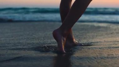 Gün batımında bir kız kumsalda yalınayak yürüyor, kumda ayak izleri ve suda yansımalar bırakıyor. Sahilde kadın bacaklarına yakın çekim. Sakinlik ve sükunet. Yaz tatilindeki turist