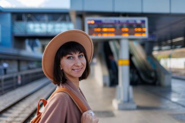 Trenle yolculuk. Paltolu ve şapkalı genç bir kadın istasyonda duruyor ve tren saatine bakıyor. macera ve seyahat.