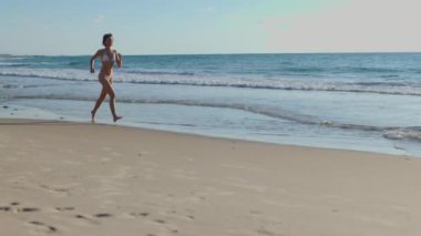 Beyaz mayo giymiş genç bir kadın kumlu sahil boyunca koşuyor. Kız sahilde koşuyor ve tatilin tadını çıkarıyor..