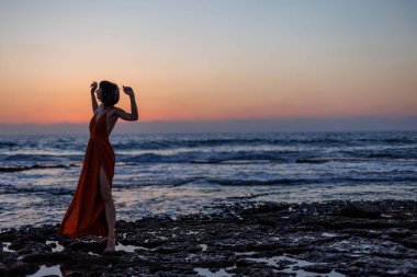 Gün batımında deniz kenarında poz veren kırmızı uzun elbiseli güzel, şehvetli bir kadın. Güzel gün batımı. Havada uçan bir kumaş şeridi..