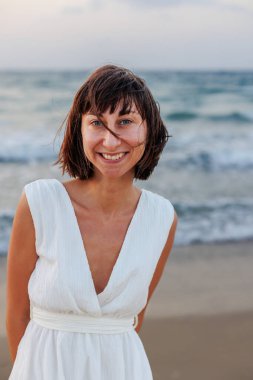 Beyaz elbiseli güzel, gülümseyen bir esmer, denizin arka planında kameraya mutlu bir şekilde bakar. Genç bir kızın portresi.