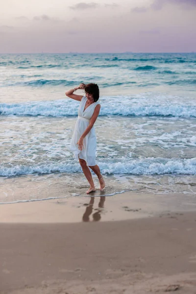 Happy traveler girl in a white summer dress enjoying a tropical paradise beach with turquoise sea. Happy young woman in a beautiful white dress walks along the beach during sunset.