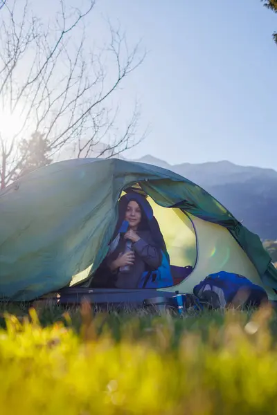 camping and trekking with children. happy smiling child sitting in a tent against a background of mountains holding a bottle of water. portrait of a child in a tent. children's outdoor recreation.