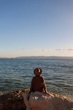 Arkadan bak. Paltolu ve şapkalı genç bir kız yüksek bir taşın üzerinde oturuyor ve mavi denizin ve gökyüzünün güzel manzarasına bakıyor. Kıyı şeridi.