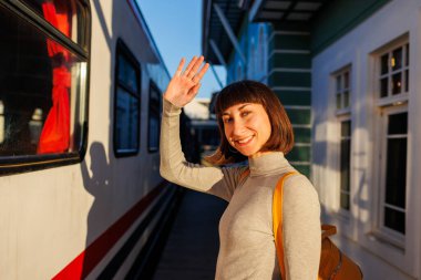 Genç gezgin platformda duruyor ve trendeki birine el sallayarak veda ediyor. Gitmeden önce hoşça kal de. seyahat ve macera.