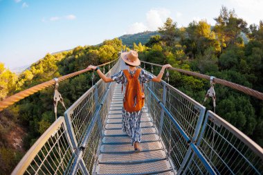 Sırt çantalı bir kız. Bir nehrin üzerindeki asma köprüde yürüyen genç bir kadının açık hava portresi. Seyahat ve macera
