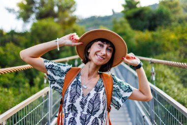 Sırt çantalı bir kız. Bir nehrin üzerindeki asma köprüde yürüyen genç bir kadının açık hava portresi. Seyahat ve macera