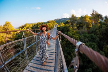 Sırt çantalı bir kız. Şapkalı ve sırt çantalı genç bir kadın nehrin üzerindeki asma köprü boyunca yürüyor. Seyahat ve macera.