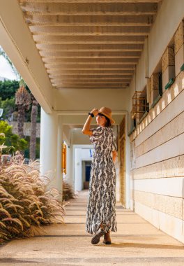 Şapka takan güzel bir bayanın tam boy fotoğrafı. Sokakta yürümek, güneşli hava, hafta sonu. Elbiseli ve şapkalı genç bir kız sokakta yürüyor..