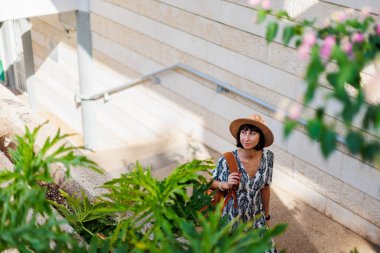 Elbiseli ve sırt çantalı bir kızın portresi. Sırt çantalı genç bir kız ağaçların arasında sokakta yürüyor. Şapkalı kız..