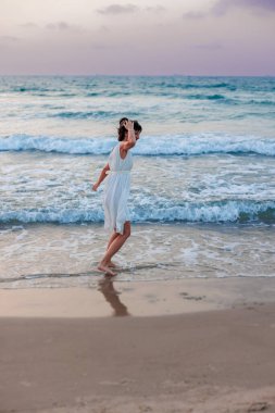 Beyaz yazlık elbiseli mutlu genç kız gün batımında sahilde yürüyor. Kumsalda dans eden kız..