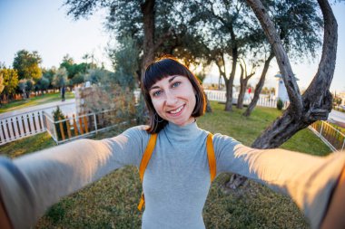 Sırt çantalı, selfie çeken ve yaz parkında yürürken kameraya bakan pozitif bir kız resmi. Kız seyahat eder.