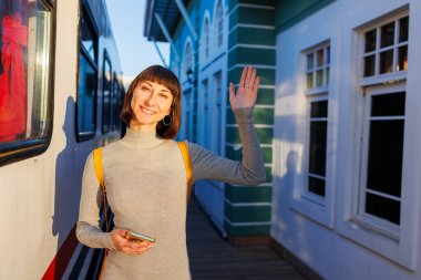 Sırt çantalı genç bir gezgin platformda duruyor ve elinde telefon olan bir kızla vedalaşıyor. Gitmeden önce hoşça kal de. seyahat ve macera.