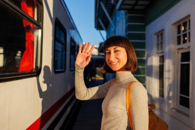 Genç gezgin platformda duruyor ve trendeki birine el sallayarak veda ediyor. Gitmeden önce hoşça kal de. seyahat ve macera.