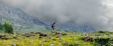 Dağlarda yürüyüş. Sırt çantalı küçük bir çocuk dağların ve bulutların ardında bir patika boyunca yürüyor. Hafta sonu yürüyüş için aktif sağlıklı yaşam tarzı. Seyahat kavramı